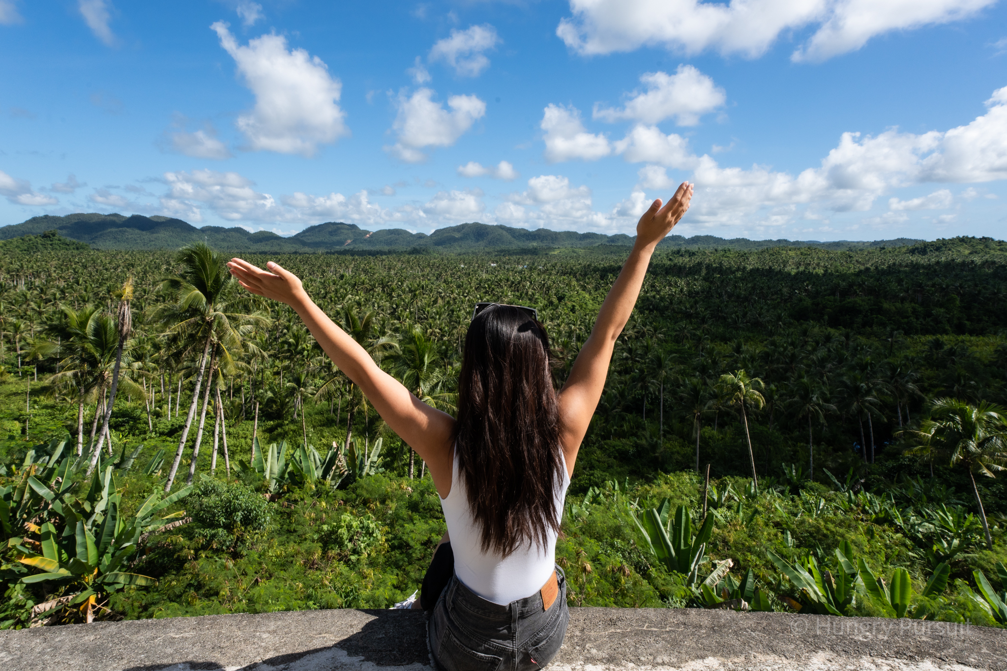 Girl sitting on the Coconut Road ledge in Siargao, hands raised in the air, embracing the island's beauty.