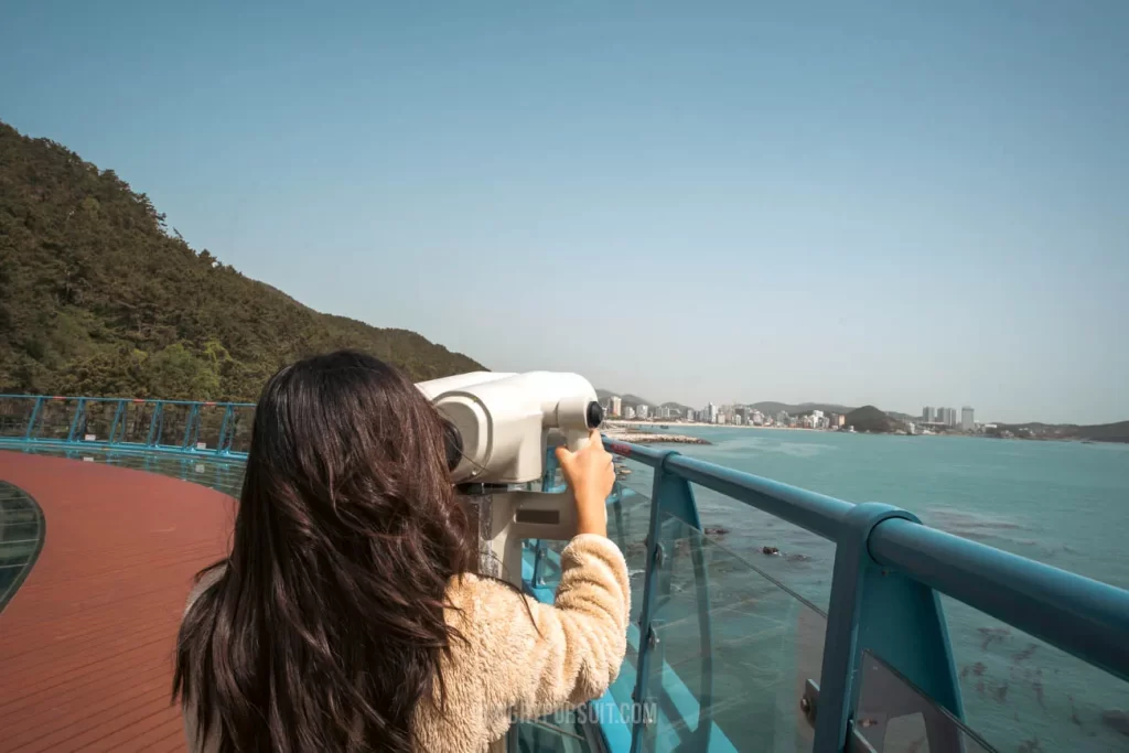 a woman at the Cheongsapo Daritdol Observatory looking through binoculars at Haeundae Blueline Park